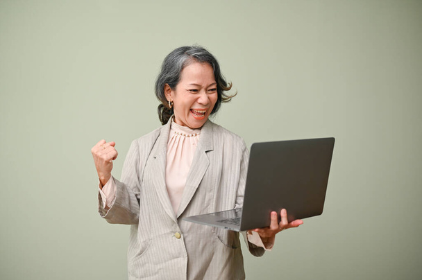 Здивований і щасливий старший азіатський бізнесмен, який тримає і дивиться на свій екран ноутбука, піднімає руку, кричачи від щастя. Так, прославляючи перемогу, перемогу, успіх. - Фото, зображення