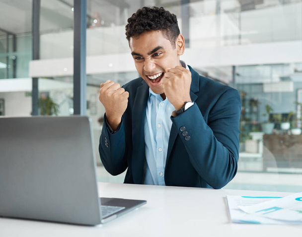 Młody biznesmen rasy mieszanej dopingujący z pasją podczas pracy nad laptopem sam w pracy. Jeden latynoski biznesmen uśmiechający się i świętujący zwycięstwo pracujący przy biurku w biurze. - Zdjęcie, obraz