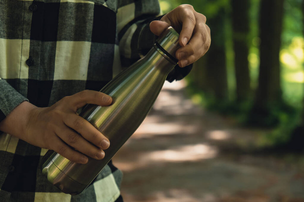Nahaufnahme einer unkenntlichen weiblichen Hand, die eine Wasserflasche hält. Mehrweg-Thermowasserflasche aus Stahl im Park. Nachhaltiger Lebensstil. Kunststofffreies, abfallfreies Leben. Grüner Umweltschutz - Foto, Bild