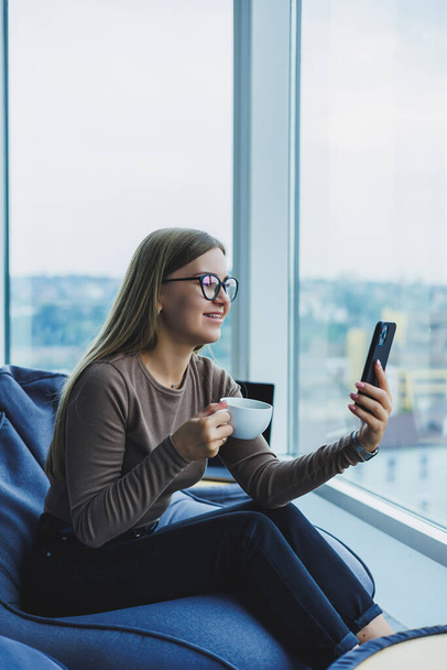 Bionda giovane donna sorridente in occhiali in abiti casual seduta su una sedia alla finestra con un telefono e bere caffè - Foto, immagini