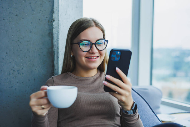 Blondi nuori hymyilevä nainen lasit rento vaatteita istuu tuolissa ikkunan vieressä puhelimen ja kahvin juominen - Valokuva, kuva