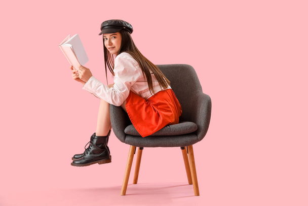 Κομψή νεαρή γυναίκα διαβάζοντας το βιβλίο στην πολυθρόνα σε ροζ φόντο - Φωτογραφία, εικόνα