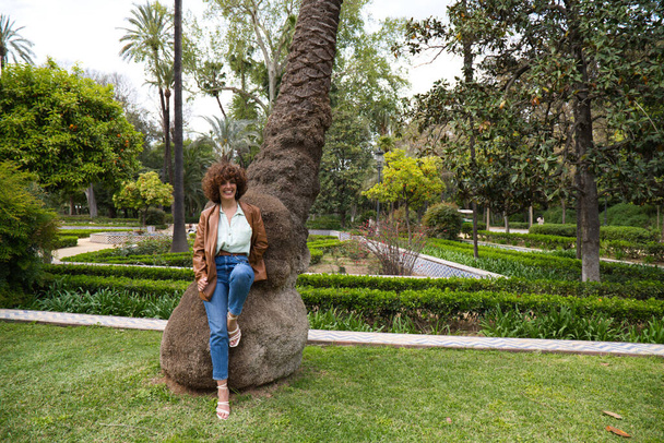 Die schöne Spanierin mit den lockigen Haaren lehnt an einem Baumstamm im Park. Die Frau ist glücklich und genießt ihren Urlaub in der Stadt. Konzept Glück, Mode und Schönheit. - Foto, Bild