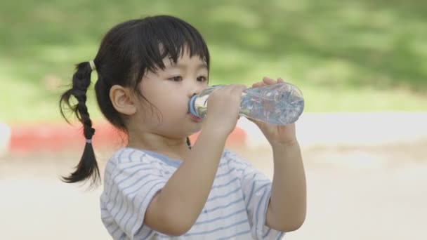 Linda niña asiática sedienta y bebiendo agua mineral después de hacer ejercicio corriendo corriendo en el parque o jardín o patio de recreo por la mañana. El desarrollo del concepto de niños pequeños. - Imágenes, Vídeo