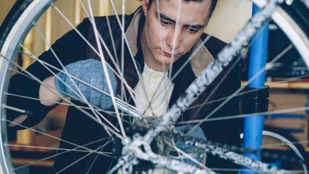 Concentrated handsome guy skilled mechanic is repairing bicycle wheel with wrenchwhile working in his workplace. Bike maintenance, people and profession concept. - Zdjęcie, obraz