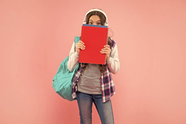 teen girl with backpack behind workbook wearing headphones on pink background, ebook. - Foto, Bild
