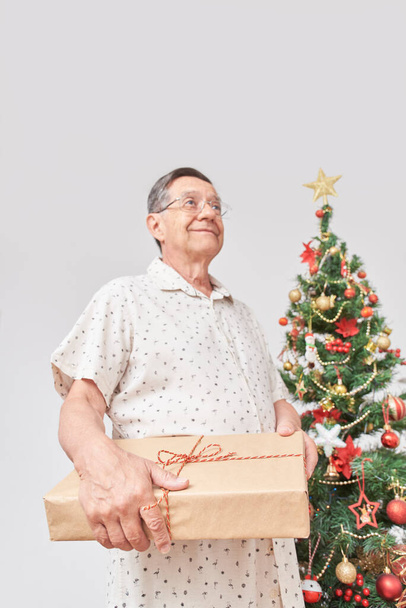 Yaşlı İspanyol adam elinde Noel hediyesi tutarken gülümsüyor. Konsept: bayramlarda hediye vermenin keyfi. Seçici odak ve kopyalama alanına sahip dikey bileşim. - Fotoğraf, Görsel
