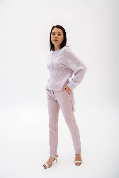 full length of trendy brunette woman in knitted sweater and pants posing with hand in pocket on white background - 写真・画像