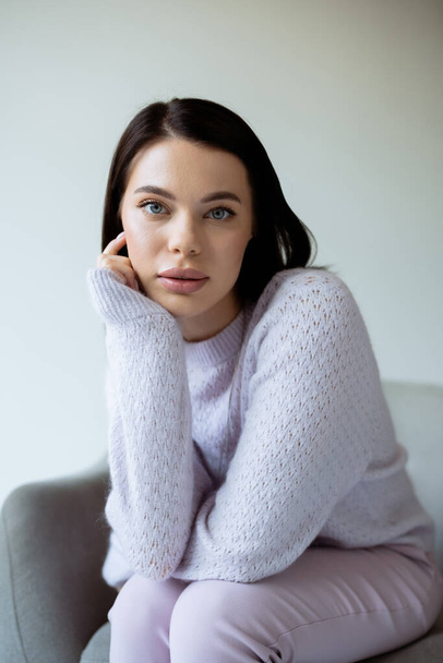 portrait of brunette woman in soft knitted sweater and natural makeup sitting isolated on grey - Foto, Bild
