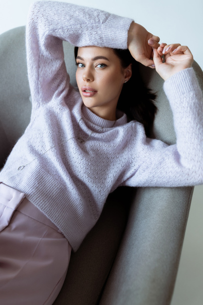 high angle view of young woman in knitted sweater sitting in armchair with hands above head isolated on grey - Photo, Image