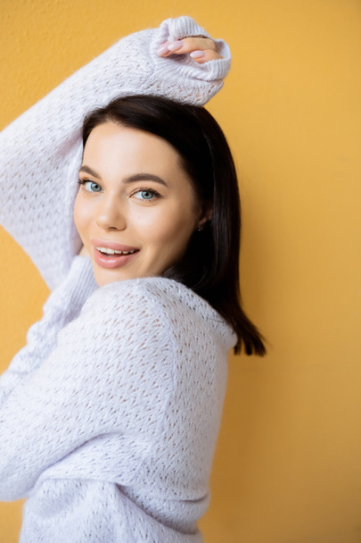 cheerful brunette woman in soft knitted sweater looking at camera while posing isolated on yellow - Фото, изображение