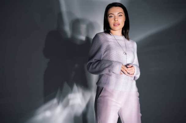 trendy woman in knitted sweater and pants standing on grey background with abstract lighting - Fotografie, Obrázek