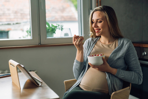 pregnant woman eats fruit salad and using digital tablet at home - Valokuva, kuva