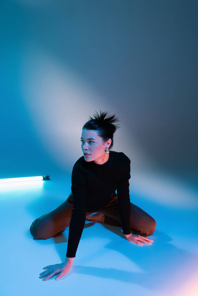 fashionable woman in black sweater sitting near fluorescent lamp and looking away on blue background - Foto, Imagem