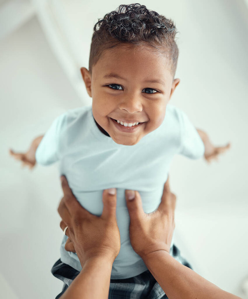 Happy little mixed race boy lying in fathers arms while looking at the camera and smiling. Loving family with father lifting cute little child son playing plane and having fun while bonding at home. - Photo, Image