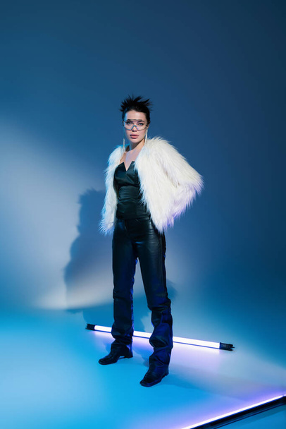full length of trendy woman in black leather pants and white furry jacket standing near fluorescent lamp on blue background - Fotoğraf, Görsel