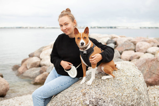 Woman with basenji dog sitting and resting outdoors on the beach. Doggy feeling happy spending time with owner in park near the sea, lifestyle photo - Фото, изображение