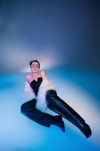 seductive model in black leather pants and white fluffy jacket lying on blue background - Foto, immagini