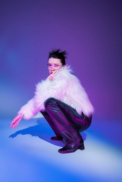 full length of brunette model in black leather pants and white furry jacket looking at camera on purple background - Photo, Image