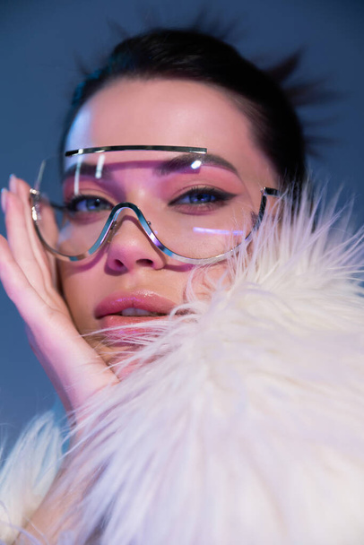 portrait of young woman in transparent sunglasses and white faux fur jacket looking at camera on blue background - Photo, Image