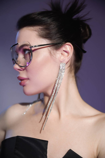 young brunette woman in shiny earrings and fashionable sunglasses looking away on purple background - Fotó, kép