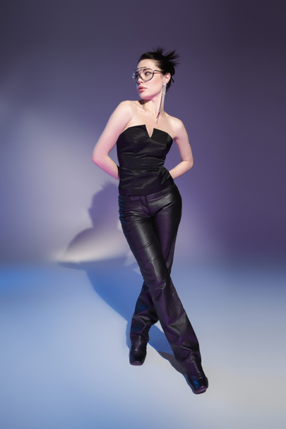 full length of glamour woman in leather pants and black corset posing with hands behind back on purple background - Photo, Image