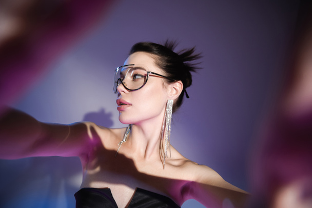 brunette woman in trendy sunglasses and shiny earrings looking away on blurred purple background - Foto, afbeelding