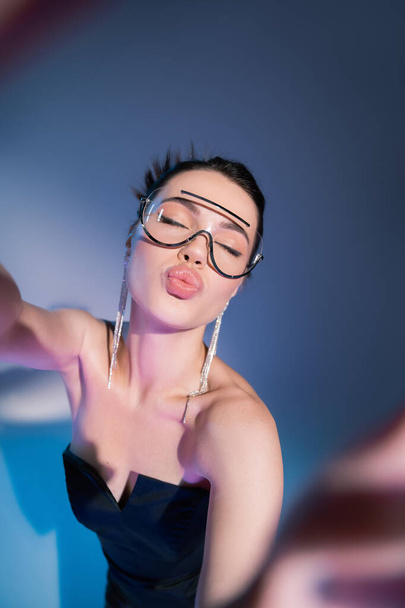 top view of young woman in transparent trendy eyeglasses pouting lips on blue background - Fotó, kép