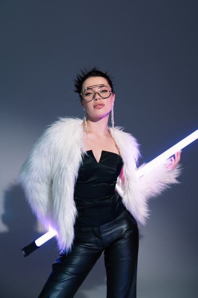 trendy woman in black corset and white faux fur jacket posing with fluorescent lamp on grey background - Photo, Image