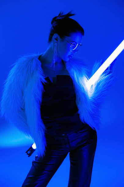 young woman in black corset and furry jacket posing with fluorescent lamp on bright blue background - Foto, afbeelding