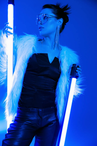 Low angle view of trendy woman in sunglasses and faux fur jacket holding lamps on blue background  - Foto, Imagem