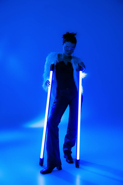 Полная длина модной женщины в искусственной меховой куртке держа лампы на синем фоне  - Фото, изображение