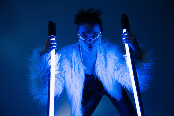 Trendy young woman in furry jacket and sunglasses holding lamps on navy blue background  - Fotó, kép