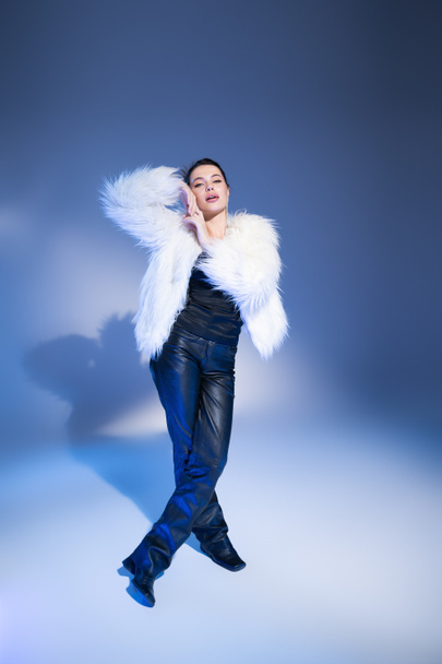 Full length of trendy brunette model in faux fur jacket posing on blue background - Photo, image