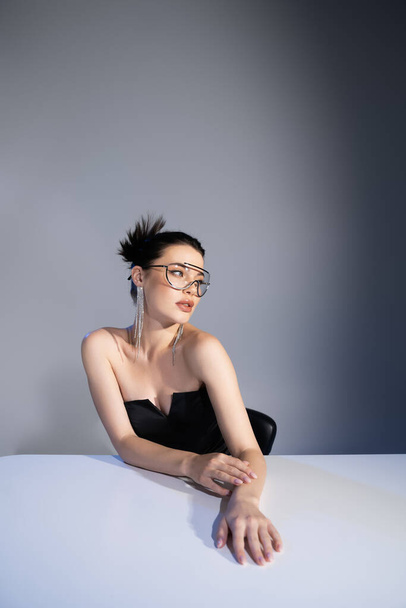 Trendy model in sunglasses and corset posing near table on grey background  - Photo, Image