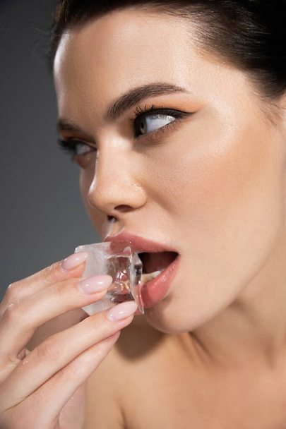Portrait of brunette woman with makeup holding ice cube near lips isolated on grey  - Photo, Image