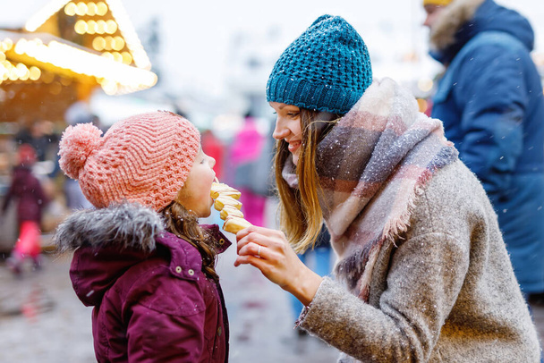 Young mother and daughter eating white chocolate covered fruits on skewer on traditional German Christmas market. Happy girl and woman on traditional family market in Germany, Munich during snowy day - Valokuva, kuva