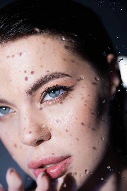 Brunette model with visage touching wet glass on grey background  - Foto, imagen