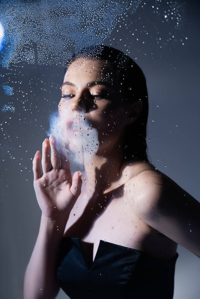 Mulher morena elegante respirando em vidro molhado em fundo cinza  - Foto, Imagem