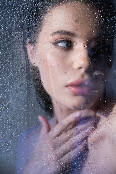 Young brunette woman touching neck behind wet glass on grey background  - Foto, Imagem
