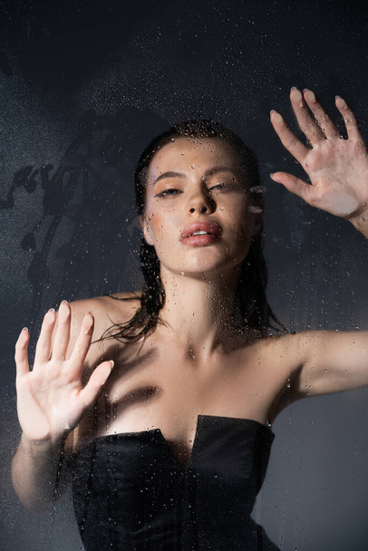 Stylish model in black corset standing behind wet glass and looking at camera on grey background  - Фото, зображення