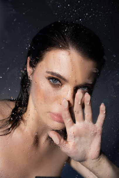 Young brunette woman with makeup touching wet glass on grey background  - Φωτογραφία, εικόνα