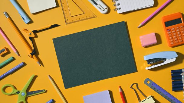 Flat lay empty green paper surrounded by various school supplies on yellow background. - 写真・画像