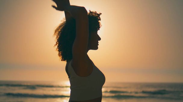 Африканская американская спортивная девушка согревает тело перед ярким восходом солнца вблизи. Силуэт атлетичной женщины изгиб тела с руками за головой стоя пляж. Тонкая спортсменка, растянувшаяся на летней природе. - Фото, изображение