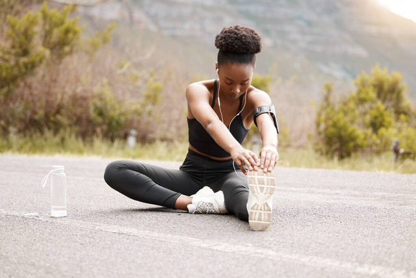 One african american female athlete with an afro listening to music on her earphones while exercising outdoors in nature. Dedicated black woman smiling while warming up before a workout outside. - Photo, Image