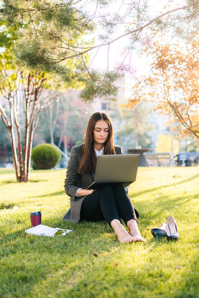 Расслабленная босоногая деловая женщина, работающая за ноутбуком, сидящая на зеленой траве в парке. Летом. Интернет-связь, беспроводное соединение - Фото, изображение