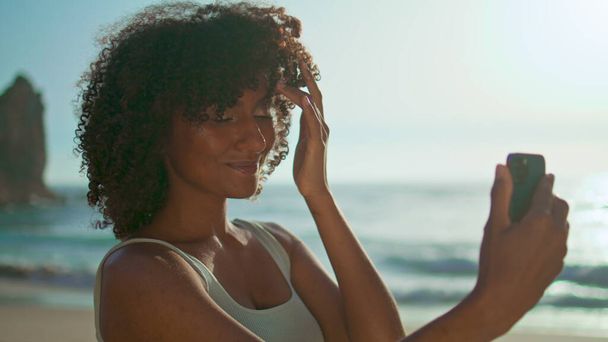 Menina sorridente despreocupado posando no smartphone fazendo selfie na praia nascer do sol closeup. Retrato de mulher americana africana atraente tirando auto foto na câmera móvel ao ar livre. Conceito de tecnologia na vida - Foto, Imagem
