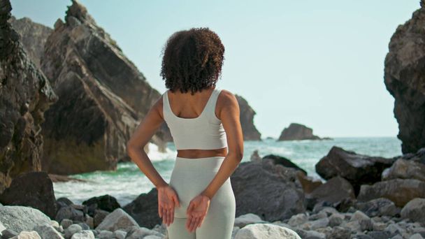 Relaxed african american yogi girl standing with Namaste hands behind back on Ursa rocky seashore. Unknown sporty woman stretching arms making yoga asana on nature. Harmony energy outdoors concept. - Photo, Image