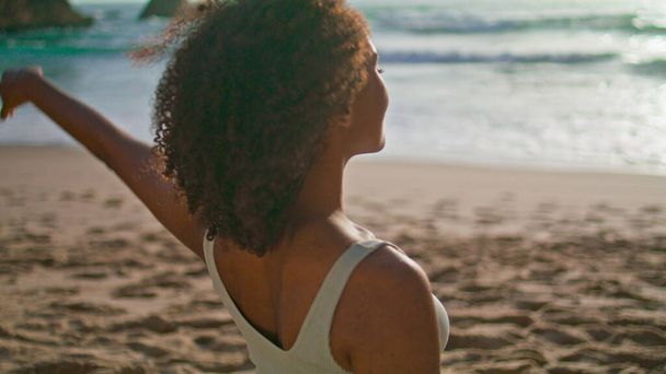 Vue de dos afro-américaine fille étirant le cou sur la plage de sable gros plan. Femme détendue et insouciante tenant la tête avec la main faisant des exercices de fitness au lever du soleil. Slim sportive faire échauffement avant l'entraînement - Photo, image