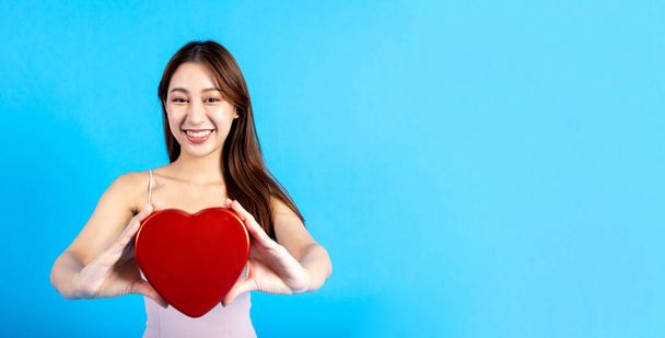 Happy smiling Asian female stylish woman holding a heart shape object in blue background. Love and Valentines day concept. Half body length studio with copy space. - Foto, immagini